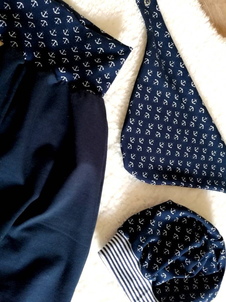 May include: Navy blue fabric items with a white anchor pattern. The items include a pair of pants, a bib, and a beanie. The beanie has a white and navy blue striped cuff. The items are on a white, fluffy surface.