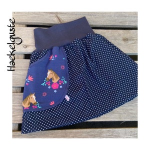 Könnte beinhalten: Ein marineblauer Rock mit weißen Punkten und einer großen Tasche auf der Vorderseite. Die Tasche hat einen blauen Hintergrund mit einem Pferd und einem Blumenmuster.