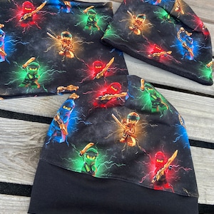 Könnte beinhalten: Schwarze Strickmütze und Halstuch mit einem sich wiederholenden Muster von Ninjas in Rot, Grün und Blau mit Blitzen auf schwarzem Hintergrund.