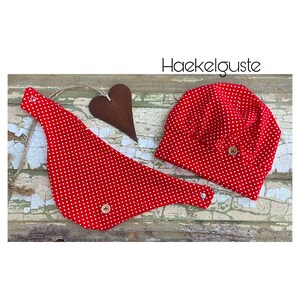 Könnte beinhalten: Rotes Baby-Mützen- und Lätzchen-Set mit weißen Punkten und Holz-Knöpfen. Das Set liegt auf einem rustikalen Holz-Hintergrund mit einem herzförmigen Stück Metall.