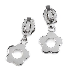 Könnte beinhalten: Zwei silberne Reißverschlussanhänger mit blütenförmigen Anhängern. Die Reißverschlussanhänger sind aus glänzendem Metall gefertigt und haben ein klassisches Design. Die Blumenanhänger haben ein einfaches, fünfblättriges Design mit einem kreisförmigen Zentrum.