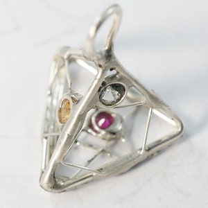 Könnte beinhalten: Ein silberner Anhänger in Form einer Pyramide mit einem Drahtkäfig-Design. Der Käfig enthält drei Edelsteine: einen roten Rubin, einen klaren Diamanten und einen gelben Saphir.