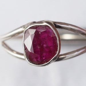 K&ouml;nnte beinhalten: Ein silberner Ring mit einem tief roten Rubin-Edelstein, der in einer Zargenfassung gefasst ist. Der Ring hat ein schlichtes, modernes Design.