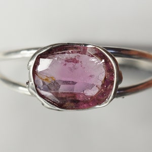 K&ouml;nnte beinhalten: Ein silberner Ring mit einem ovalen rosafarbenen Edelstein, der in einer Zargenfassung gefasst ist. Der Ring hat ein schlichtes, minimalistisches Design.