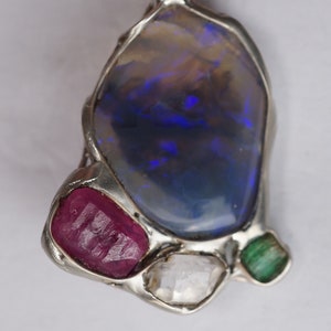 Puede incluir: Un colgante de plata con una gran piedra de ópalo azul rodeada de piedras preciosas más pequeñas: un rubí rojo, un diamante transparente y una esmeralda verde.