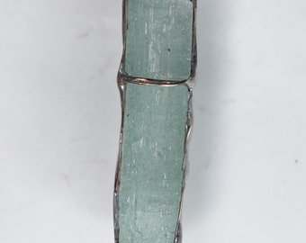 May include: A silver pendant featuring a light green aquamarine crystal, encased in a silver frame. The crystal is long and thin, with a smooth, polished surface.
