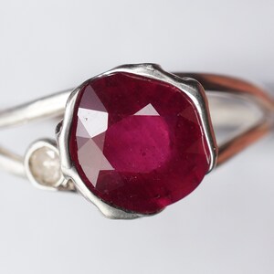 Op de afbeelding: Een zilveren ring met een grote, gefacetteerde robijn en een kleinere diamant accent. De robijn is diep rood van kleur en de ring heeft een uniek, organisch ontwerp.