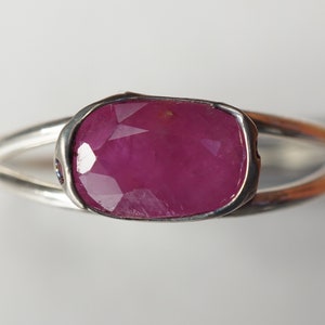May include: A silver ring with a large, faceted, pink ruby gemstone set in a bezel setting. The ring has a split shank.