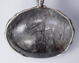 May include: A silver pendant with a clear quartz stone containing black inclusions. The stone is set in a silver bezel and the pendant has a silver bail.