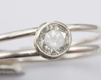 May include: A silver ring with a single, round, clear gemstone set in a bezel setting. The ring has a double band.