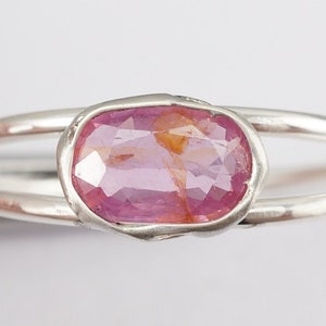 Puede incluir: Un anillo de plata con una piedra ovalada rosa engastada en un engaste de bisel. El anillo tiene una banda doble.