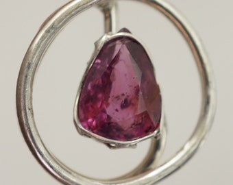 May include: A silver pendant featuring a pink gemstone set within a circular frame. The pendant hangs from a silver bail.