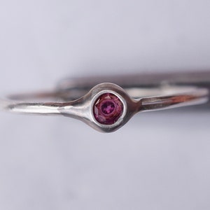 Puede incluir: Un anillo de plata con una peque&ntilde;a piedra preciosa redonda de color p&uacute;rpura engastada en el centro.