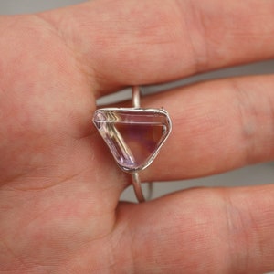 May include: A silver ring featuring a triangular amethyst gemstone. The gemstone is clear with a pale purple hue.