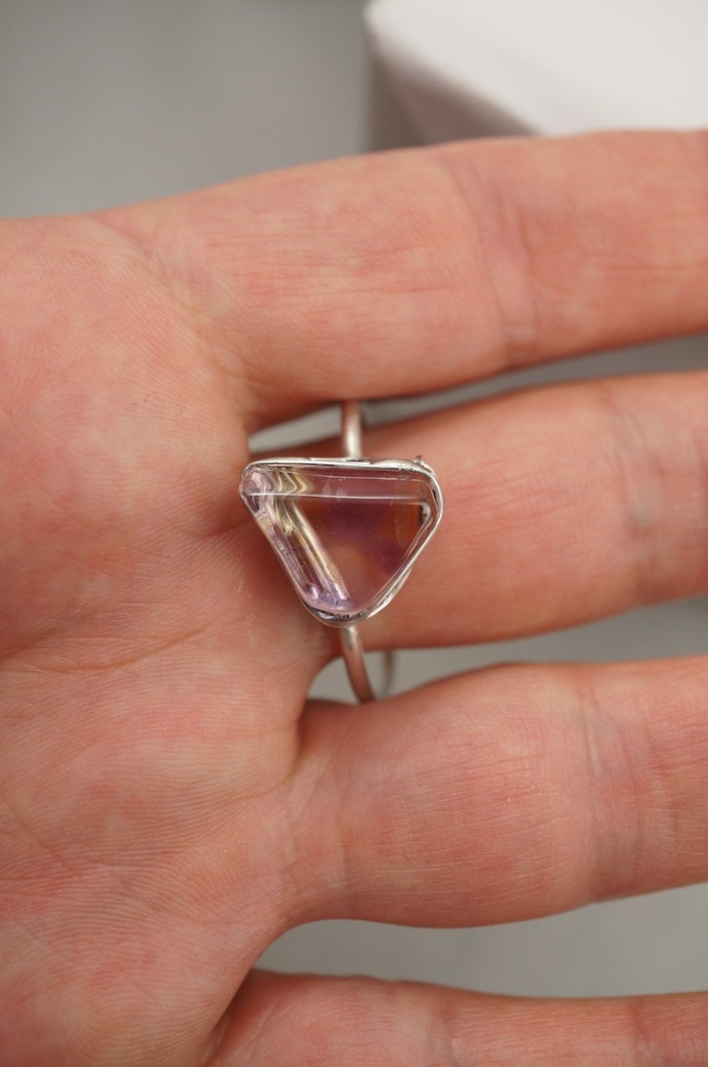 May include: A silver ring featuring a triangular amethyst gemstone. The gemstone is clear with a pale purple hue.