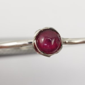 K&ouml;nnte beinhalten: Ein silberner Ring mit einem tief roten Edelstein, der in einem zarten, gedrehten Band gefasst ist.