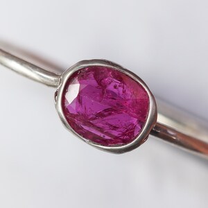 May include: A silver ring featuring a vibrant, oval-shaped, faceted, magenta gemstone. The gemstone is set in a bezel setting, and the ring band is thin and polished. The ring is displayed against a white background.