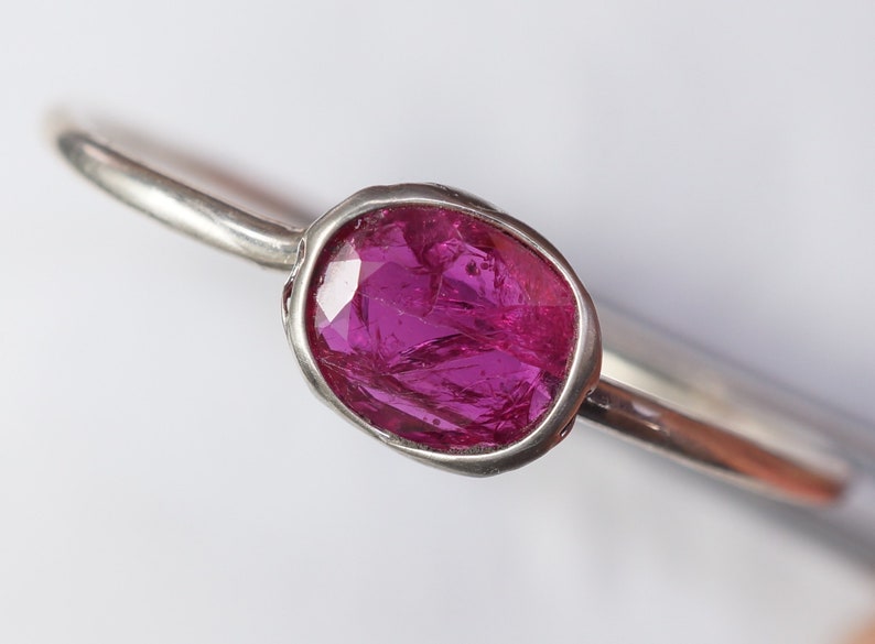 May include: A silver ring featuring a vibrant, oval-shaped, faceted, magenta gemstone. The gemstone is set in a bezel setting, and the ring band is thin and polished. The ring is displayed against a white background.