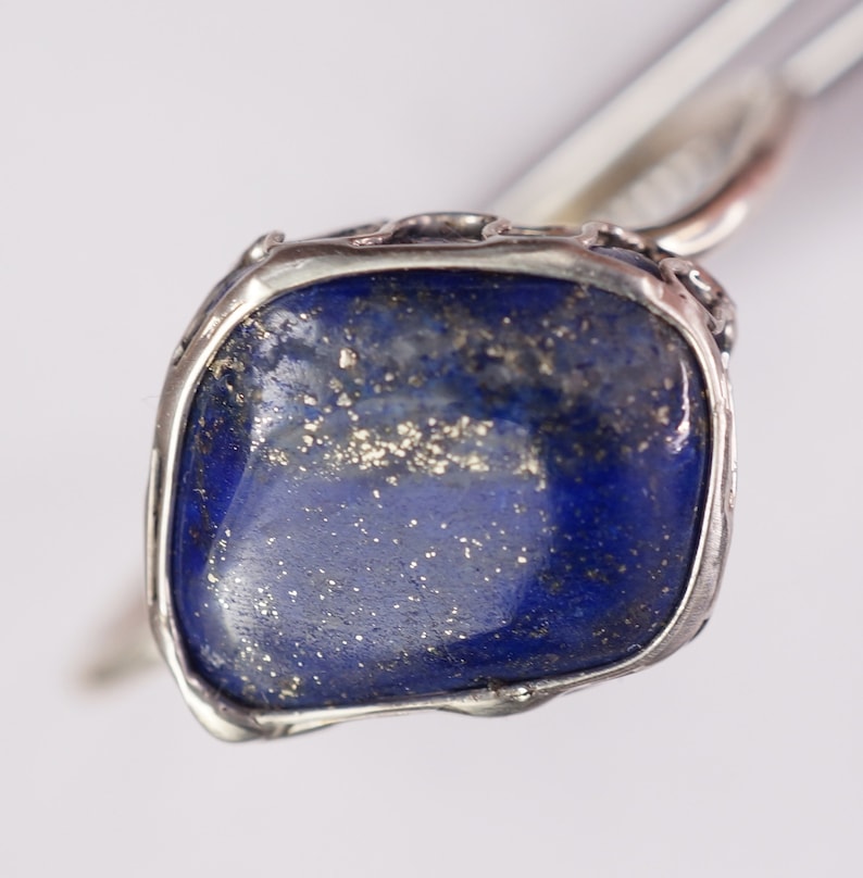 Puede incluir: Un anillo de plata con una gran piedra de lapisl&aacute;zuli azul engastada en un engaste de plata texturizado. La piedra tiene un aspecto moteado con motas doradas.