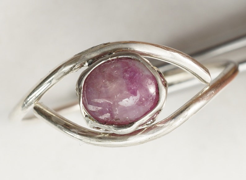 May include: A silver ring featuring a pink gemstone set in a bezel setting. The ring is designed in the shape of an eye.
