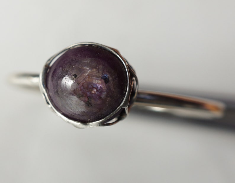 May include: A silver ring with a purple gemstone set in a wire cage. The gemstone is round and has a slightly cloudy appearance.