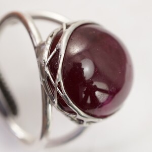 May include: A silver ring featuring a large, round, dark red gemstone set within a delicate, intricate silver cage.
