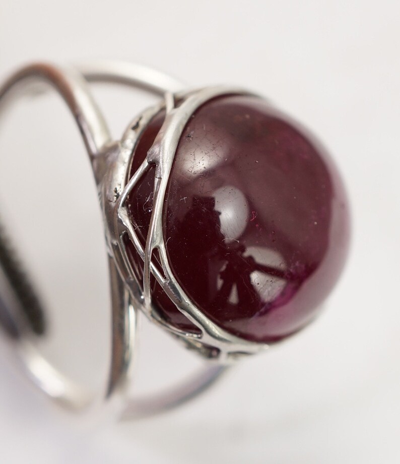 May include: A silver ring featuring a large, round, dark red gemstone set within a delicate, intricate silver cage.