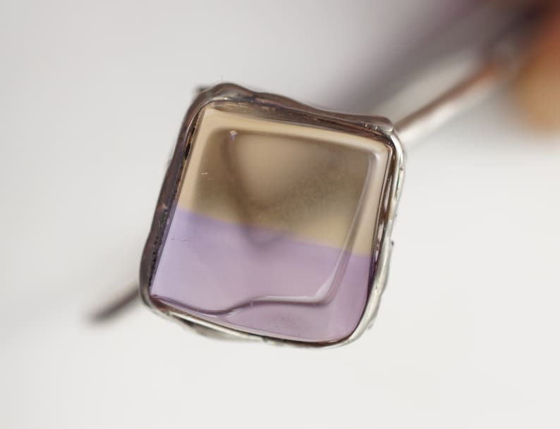 Op de afbeelding: Een zilveren ring met een vierkante, tweekleurige glazen steen. De bovenste helft van de steen is lichtbruin van kleur, en de onderste helft is lichtpaars.