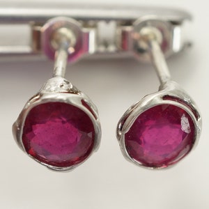 May include: A pair of silver stud earrings with a round, deep red ruby gemstone in each.