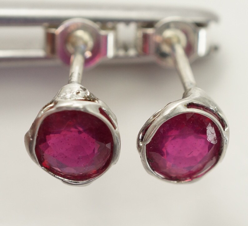 May include: A pair of silver stud earrings with a round, deep red ruby gemstone in each.