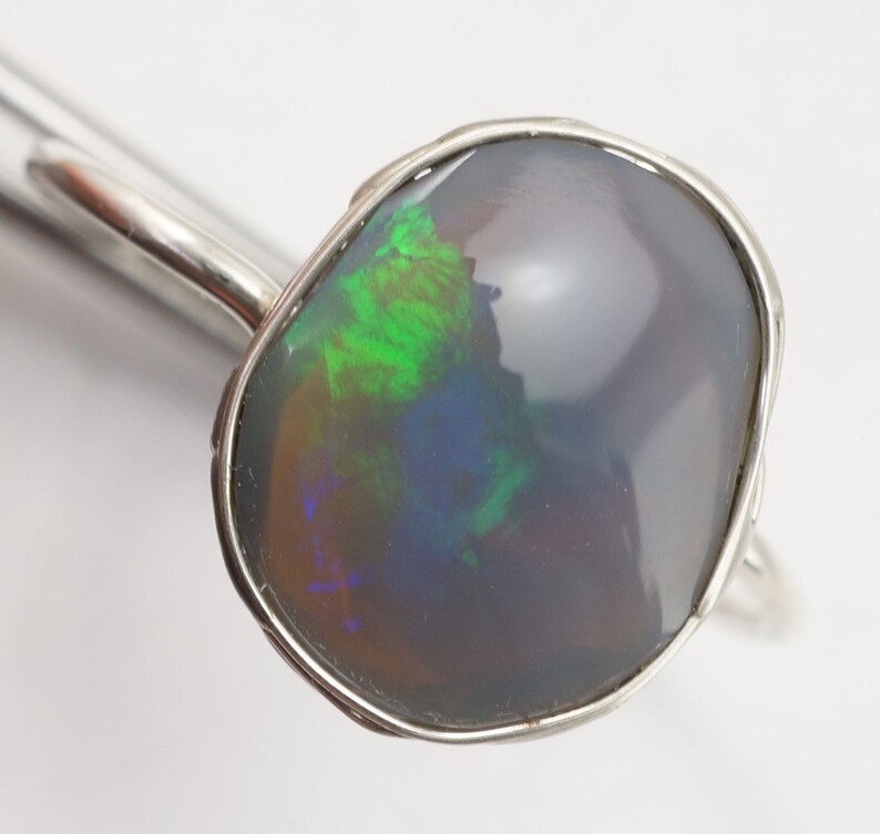 May include: A silver ring with a large, oval-shaped black opal gemstone. The opal has a green, blue, and purple iridescence.