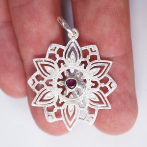 May include: A silver pendant in the shape of a mandala with a small, dark purple gemstone in the centre.