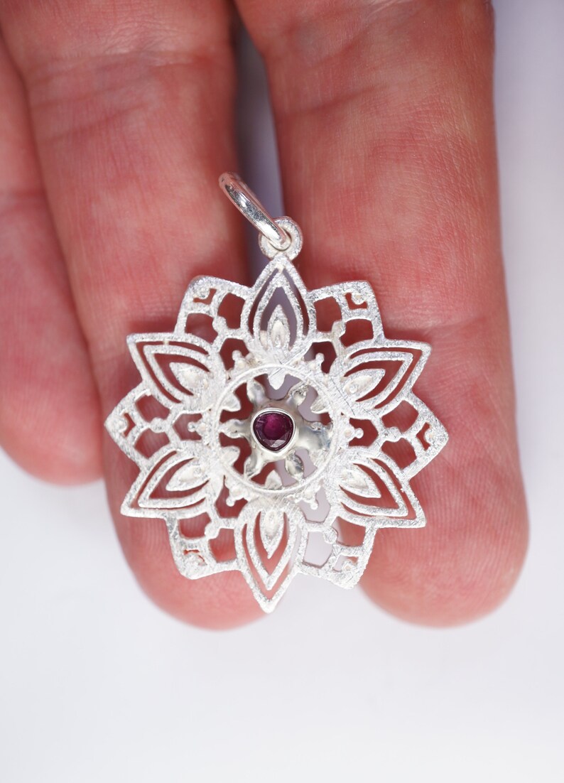 May include: A silver pendant in the shape of a mandala with a small, dark purple gemstone in the centre.