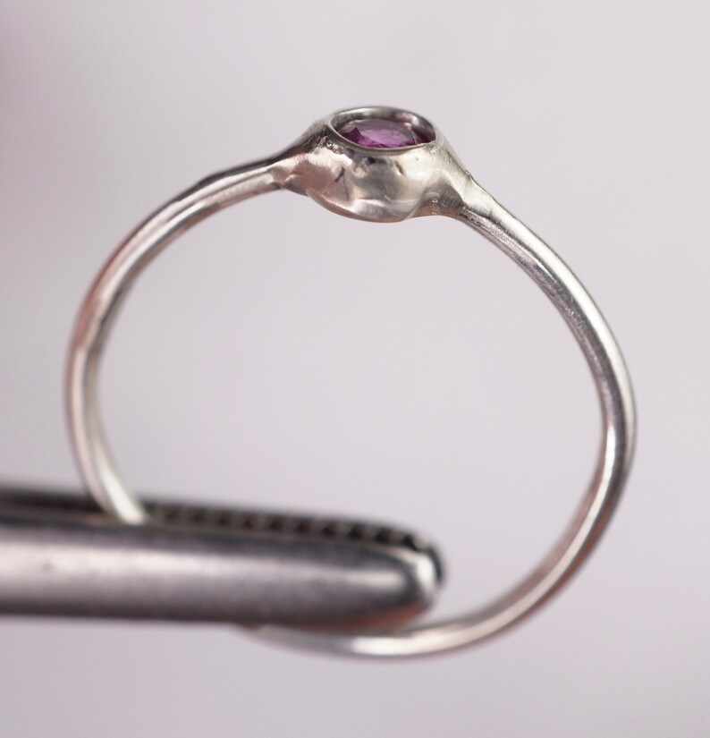 Pu&ograve; includere: Un anello d'argento con una piccola pietra preziosa rotonda e viola incastonata in una montatura a castone.