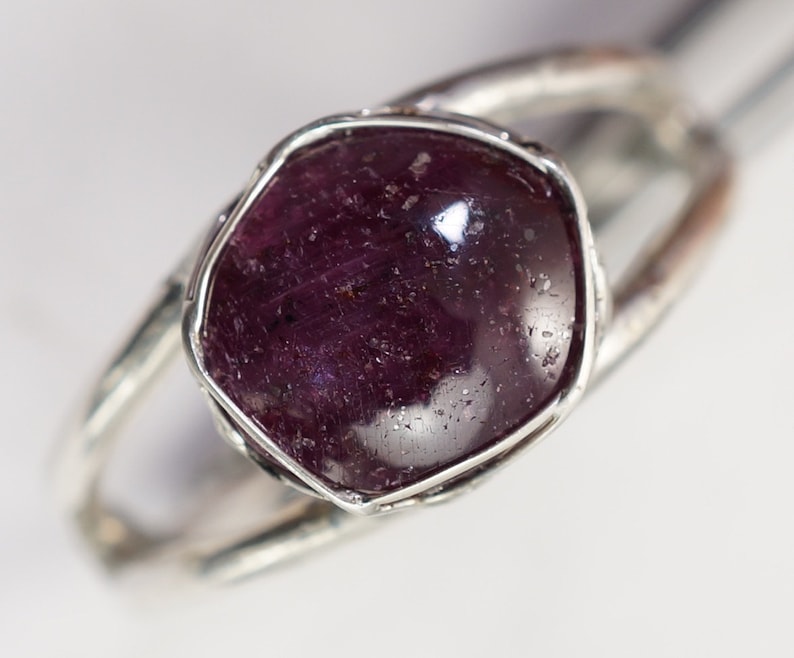 May include: A silver ring with a deep purple gemstone set in a rough, textured bezel.
