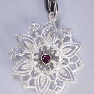 May include: A silver pendant with a delicate, intricate design featuring a small, dark purple gemstone in the centre.