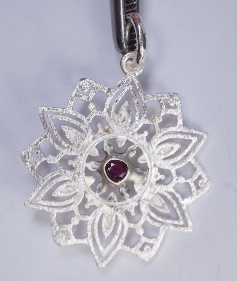 May include: A silver pendant with a delicate, intricate design featuring a small, dark purple gemstone in the centre.