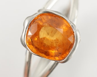 May include: A silver ring with a large, orange gemstone set in a bezel setting. The ring has a simple, modern design.