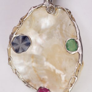 May include: A silver pendant featuring a white pearl shell, set with a blue sapphire, a green emerald, and a red ruby.