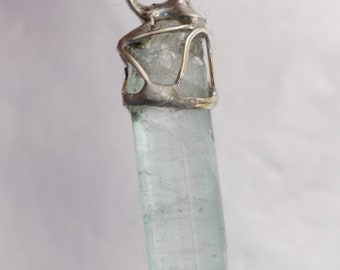 May include: A light blue aquamarine crystal pendant with a silver setting. The crystal is long and slender, with a smooth, polished surface.