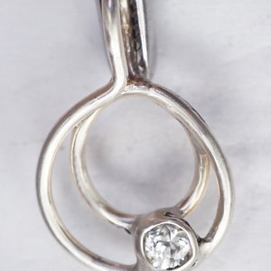 May include: A silver pendant with a clear, round gemstone. The pendant has a unique, intertwined design with a circular shape. The gemstone is set in a bezel setting, and the pendant is held by a pair of metal tweezers.