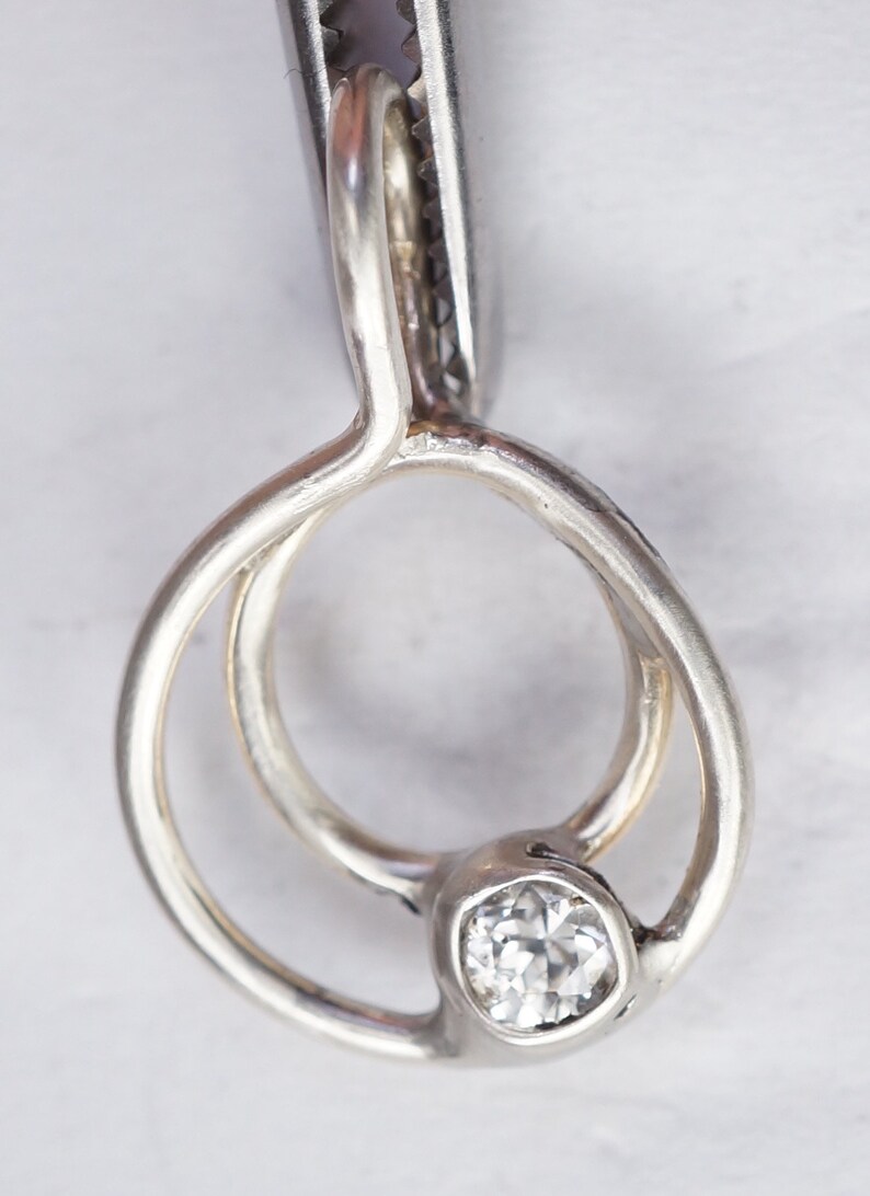 May include: A silver pendant with a clear, round gemstone. The pendant has a unique, intertwined design with a circular shape. The gemstone is set in a bezel setting, and the pendant is held by a pair of metal tweezers.