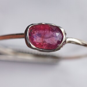 May include: A delicate silver ring with a pink oval gemstone set in a bezel setting. The ring has a simple band and is accented with small white gemstones.