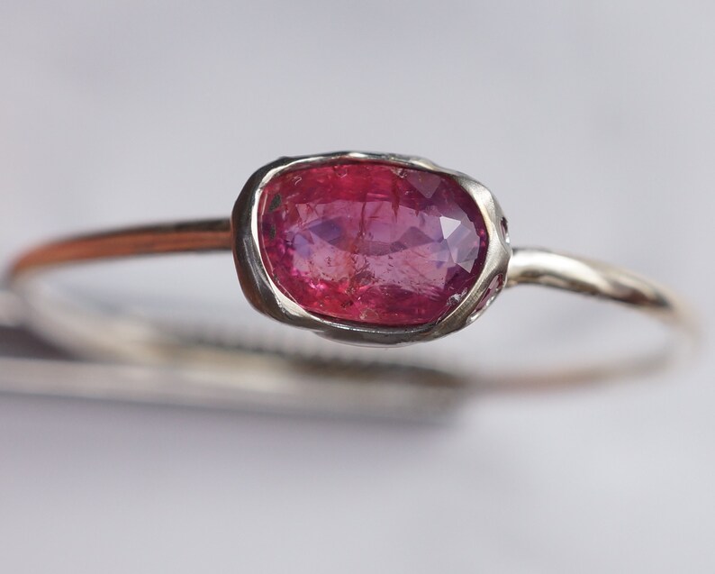 May include: A delicate silver ring with a pink oval gemstone set in a bezel setting. The ring has a simple band and is accented with small white gemstones.
