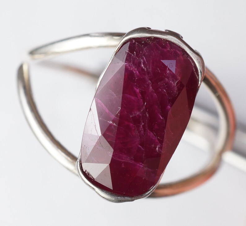 Op de afbeelding: Een zilveren ring met een grote, gefacetteerde robijn. De robijn is diep rood van kleur en heeft een licht onregelmatige vorm.
