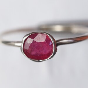 May include: A silver ring featuring a faceted ruby gemstone. The ring has a simple, delicate band and a bezel setting.