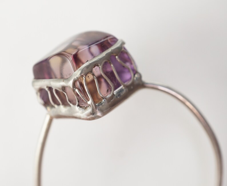 Op de afbeelding: Een zilveren ring met een grote, gefacetteerde, paarse edelsteen die in een zilveren kooi is gezet.