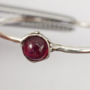 K&ouml;nnte beinhalten: Ein silberner Ring mit einem einzigen, runden, cabochon geschliffenen, dunkelroten Edelstein, der in einer einfachen, strukturierten Fassung sitzt.