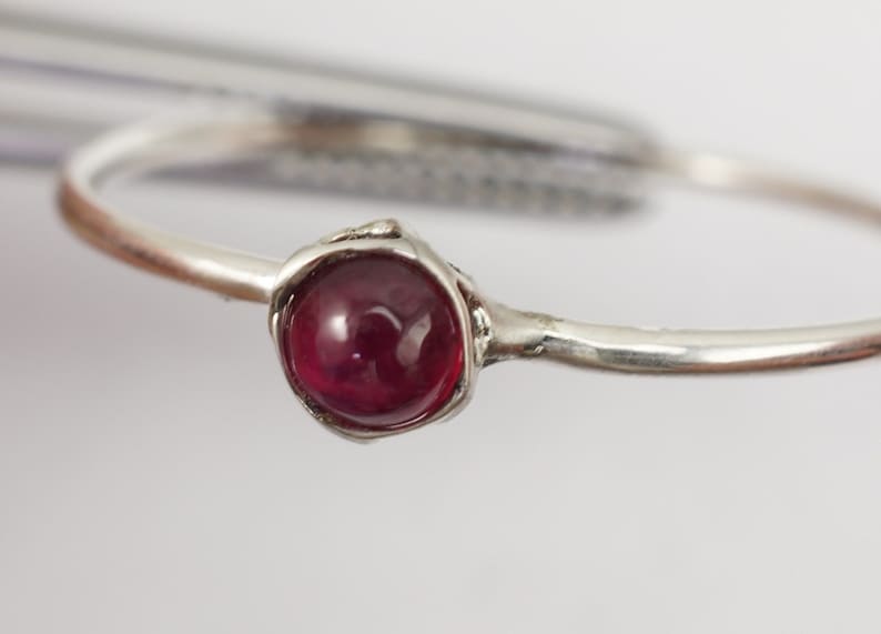 K&ouml;nnte beinhalten: Ein silberner Ring mit einem einzigen, runden, cabochon geschliffenen, dunkelroten Edelstein, der in einer einfachen, strukturierten Fassung sitzt.