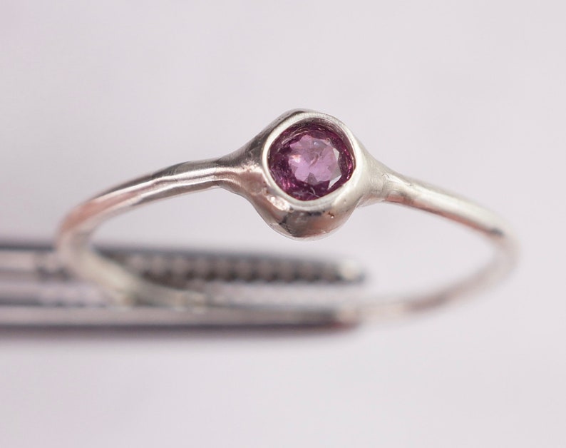 Pu&ograve; includere: Un anello d'argento con una piccola pietra preziosa rotonda di colore viola incastonata in una fascia testurizzata.